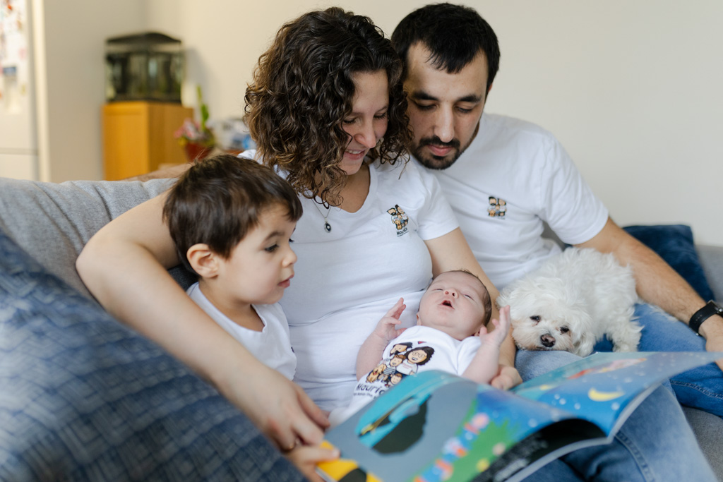 Maman tient le bébé, l’aîné regarde une histoire sur le canapé, séance naissance à domicile à Saint-Maur