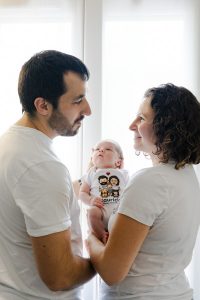 Photo en contre-jour à domicile : les parents se regardent et tiennent leur nouveau-né entre eux devant une fenêtre, séance naissance à Paris