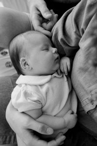 Photo rapprochée de bébé à domicile : maman tient le bébé, papa pose ses mains sur sa tête, les mains des parents illustrant la petite taille du bébé, séance naissance à Paris