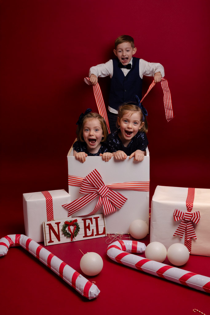 Grand frère ouvrant une boîte cadeau géante d'où sortent deux jumelles, décor de Noël rouge et blanc en studio photo à Saint-Maur