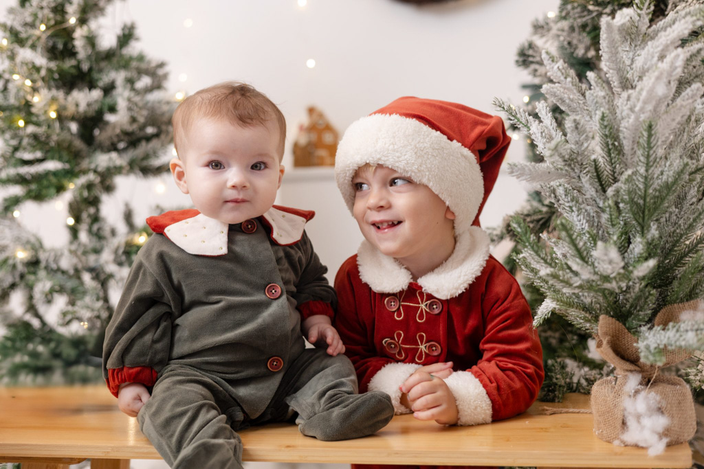 Frère et sœur en tenues de Père Noël et lutin devant un décor de Noël féerique avec sapins, guirlandes lumineuses et fausse neige.