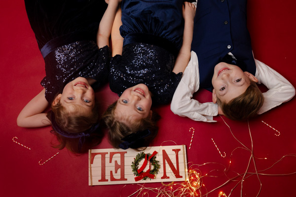 Vue du dessus des deux jumelles et de leur grand frère allongés au sol avec une plaque Noël et des décorations festives autour