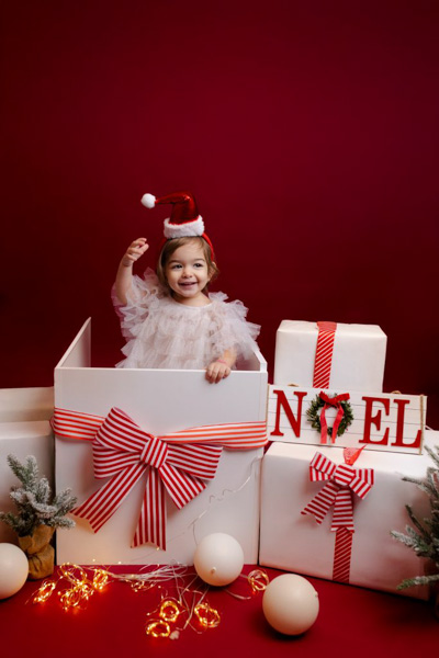 Fillette avec un bonnet de Père Noël sortant d'une grande boîte cadeau, entourée de décorations de Noël et de paquets blancs, sur fond rouge en studio photo